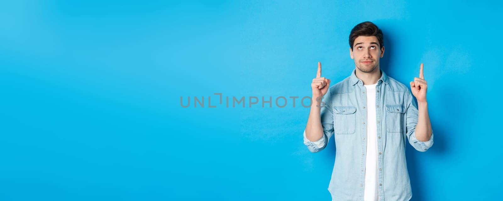 Portrait of displeased and skeptical male model pointing fingers up, looking at something unpleasant, standing against blue background.