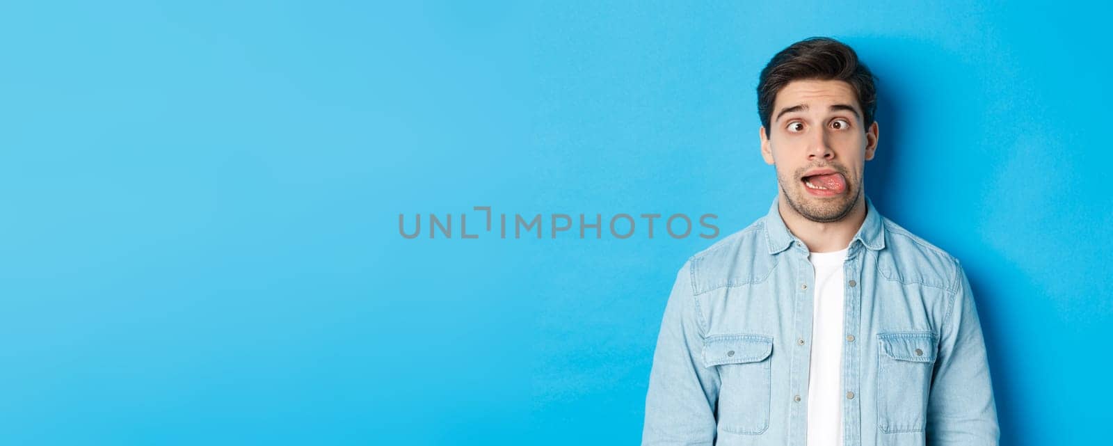 Close-up of young man making funny expressions, showing tongue and looking at camera, standing over blue background. Copy space