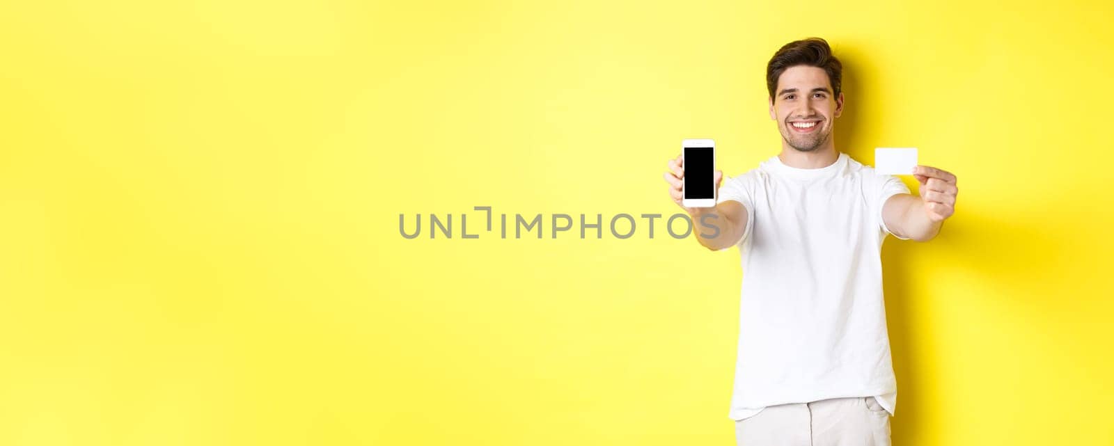 Handsome caucasian male model showing smartphone screen and credit card, concept of mobile banking and online shopping, yellow background by Benzoix