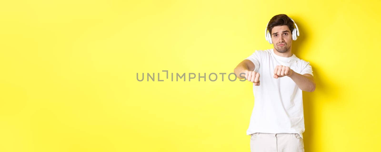 Cool guy listening music in headphones and dancing, standing in white clothes against yellow studio background.