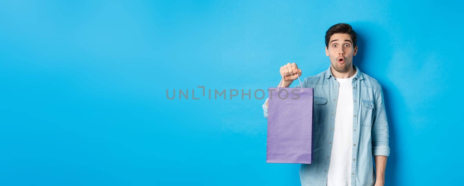 Concept of shopping, holidays and lifestyle. Handsome surprised guy holding paper bag from shop and looking amazed, standing over blue background by Benzoix