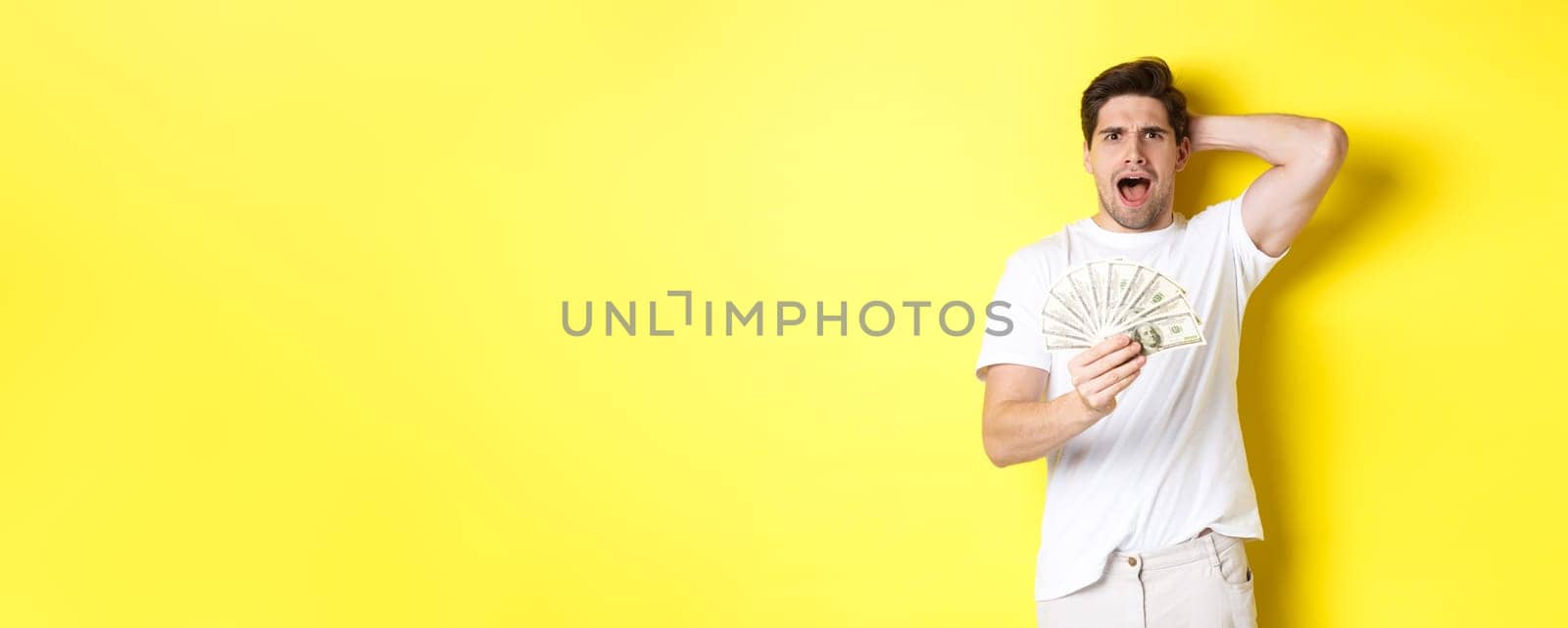 Frustrated man holding money, shouting and panicking, standing over yellow background.