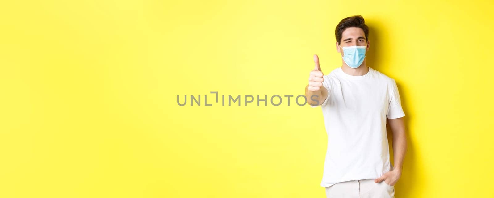 Concept of coronavirus, pandemic and social distancing. Confident young man in medical mask showing thumbs up and winking, yellow background by Benzoix