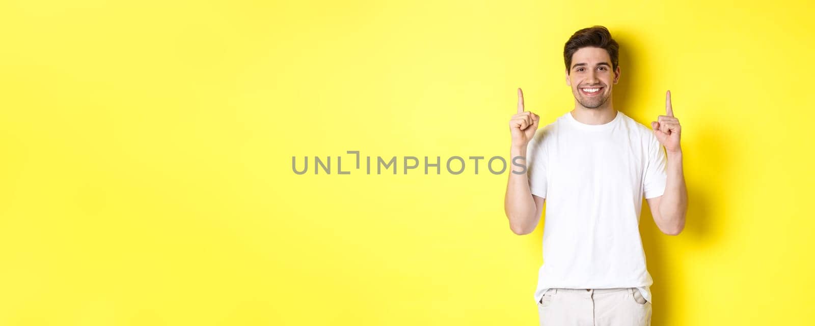 Handsome guy in white t-shirt pointing fingers up, showing shopping offers, standing over yellow background by Benzoix