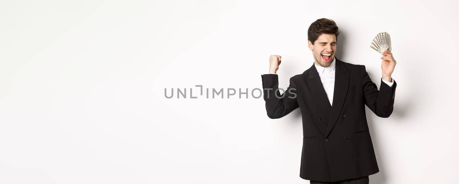 Portrait of handsome and successful businessman in suit, dancing with money, standing against white background.