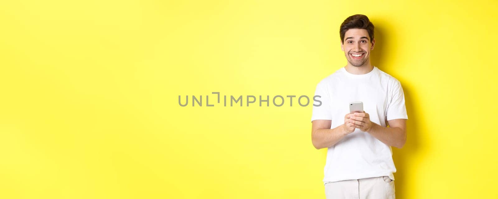 Man smiling and looking happy after reading promo offer on smartphone, standing against yellow background in white t-shirt.