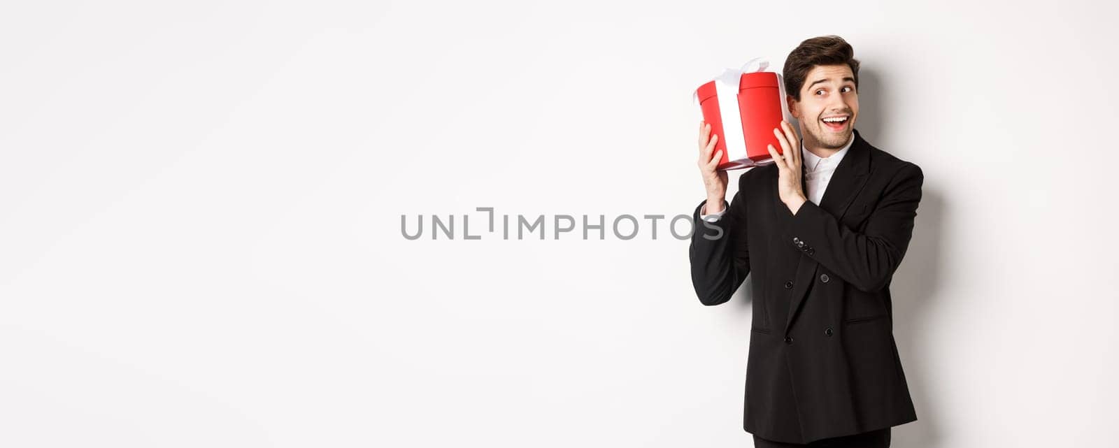 Concept of christmas holidays, celebration and lifestyle. Handsome man in black suit, shaking a box with gift, wondering whats inside, standing against white background.