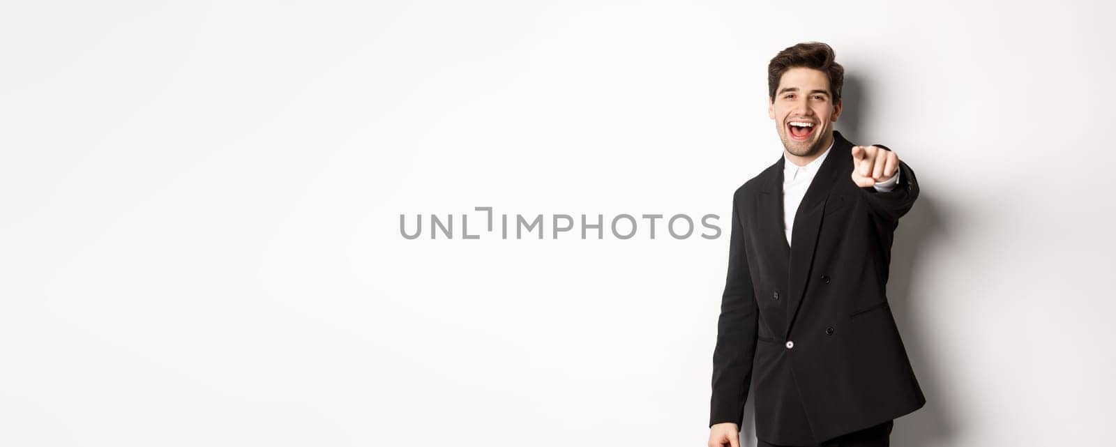 Concept of new year party, celebration and lifestyle. Portrait of handsome stylish man in black suit, smiling and pointing finger at camera, standing over white background by Benzoix