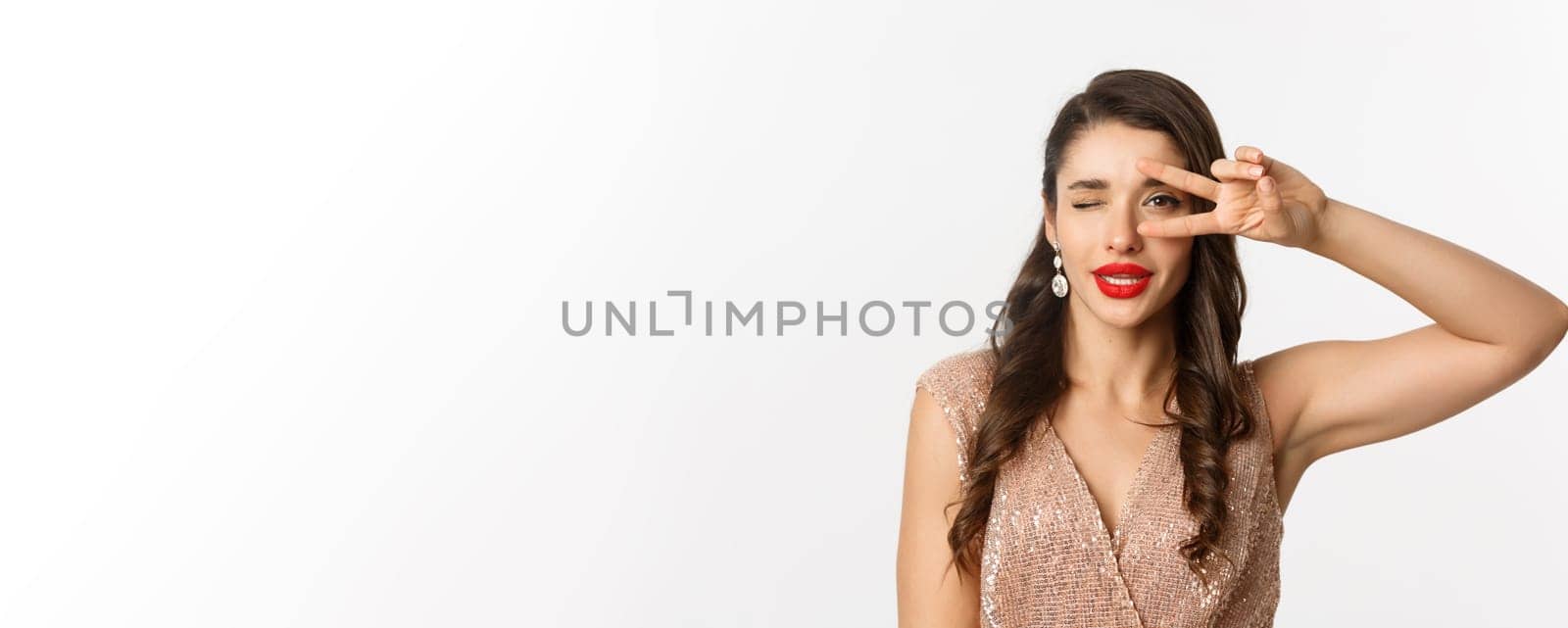 Concept of New Year celebration and winter holidays. Close-up of glamour woman in party dress, red lips, showing peace sign and winking sexy at camera, white background.