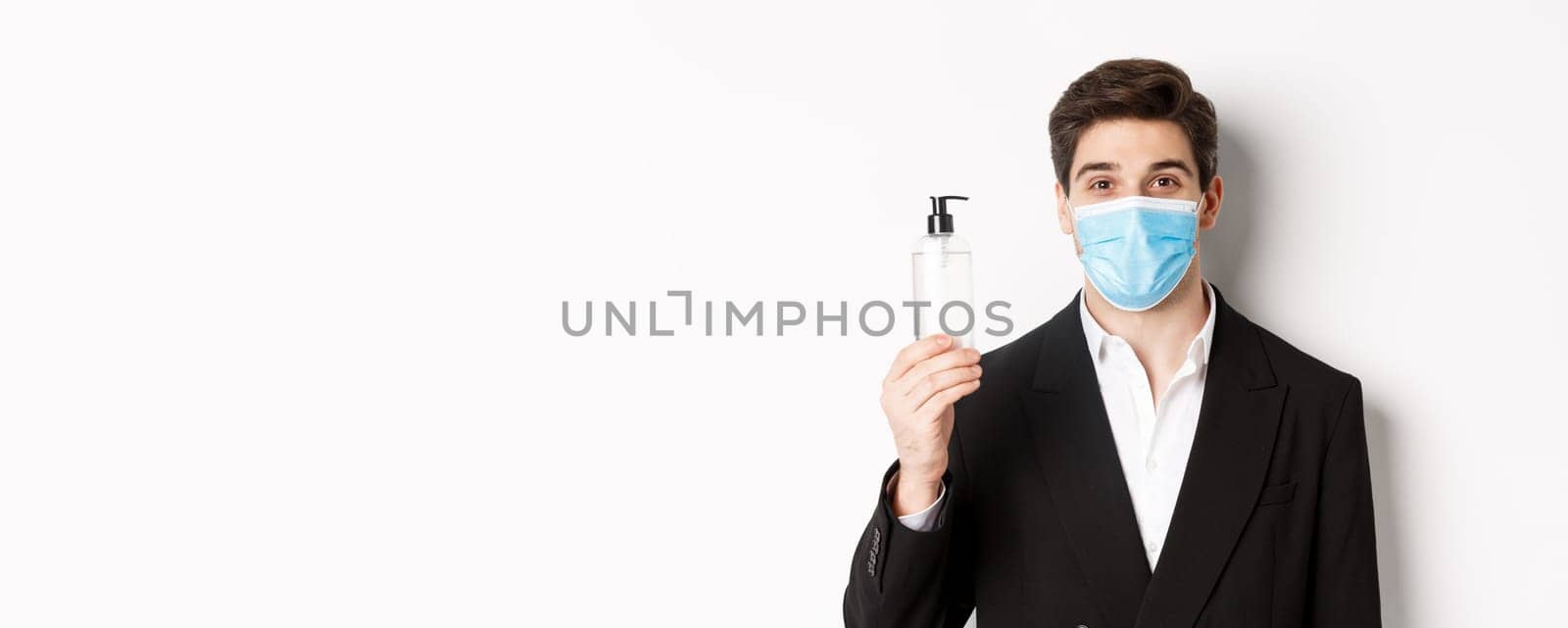 Concept of covid-19, business and social distancing. Close-up of handsome man in trendy suit and medical mask, showing hand sanitizer, standing against white background by Benzoix