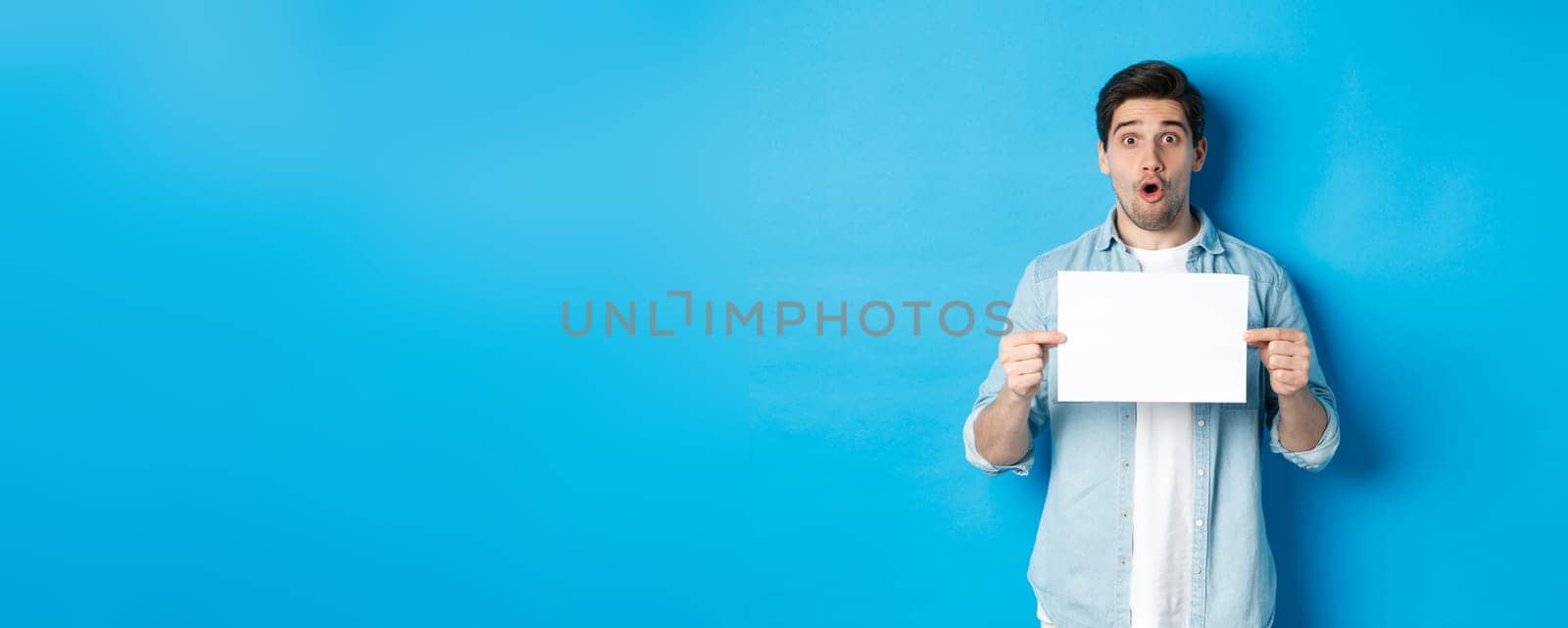 Surprised man gasping and looking impressed at camera, showing blank piece of paper for your sign, standing over blue background.