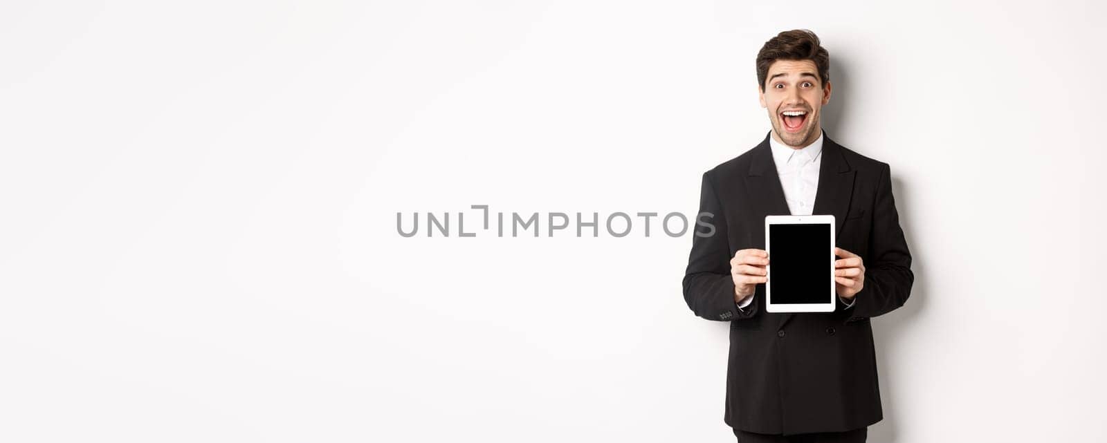 Image of attractive male entrepreneur in trendy suit, showing digital tablet screen and smiling amazed, standing over white background.
