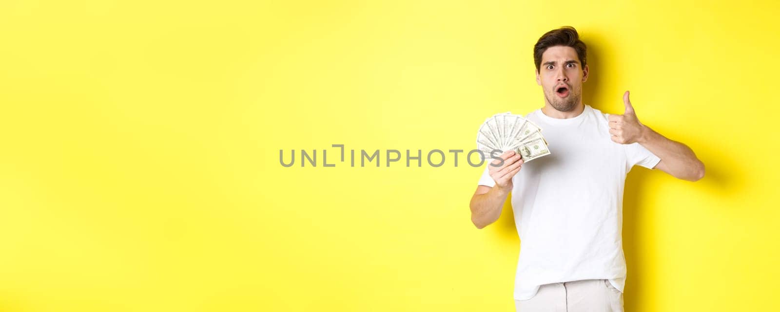 Impressed man showing thumb up, holding money credit, standing over yellow background.