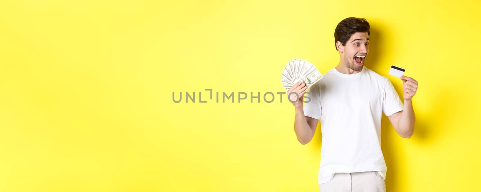 Cheerful guy looking at credit card, holding money, concept of bank credit and loans, standing over yellow background.