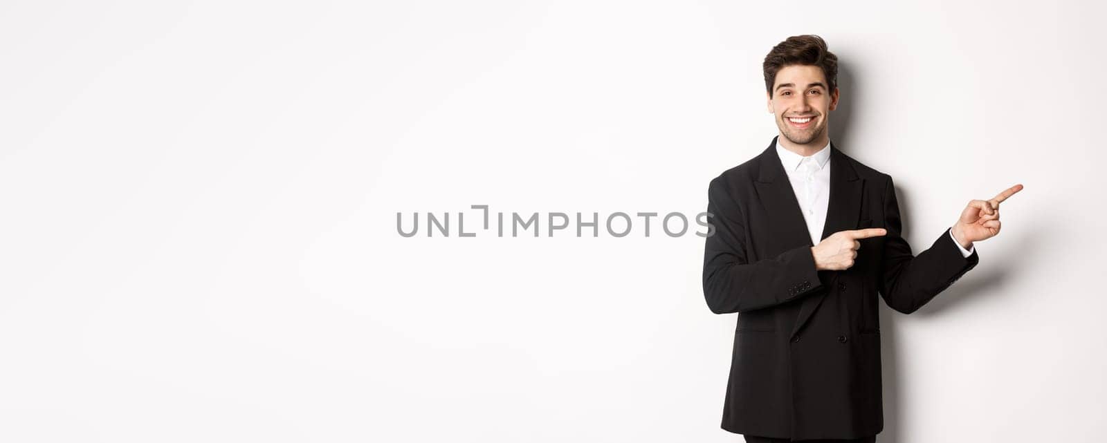 Image of attractive smiling guy dressed for new year party, pointing fingers right and showing advertisement, standing over white background.