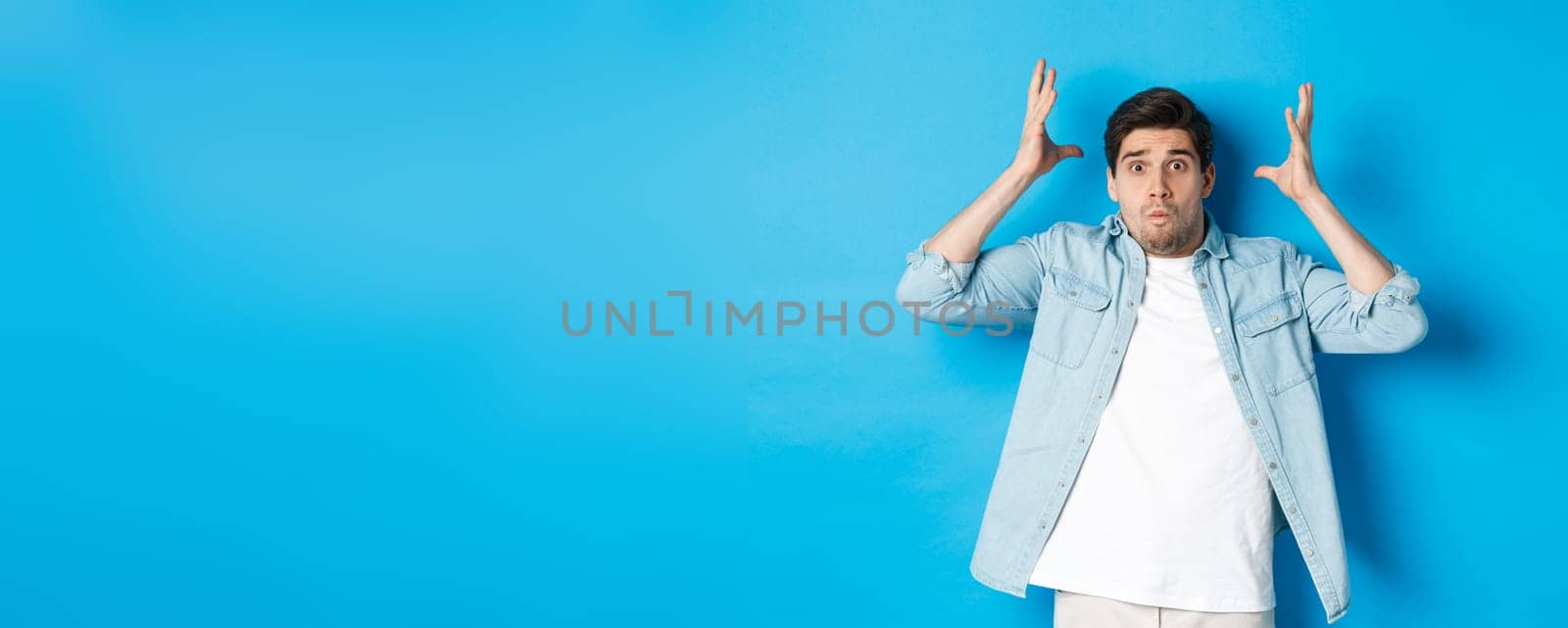 Distressed guy showing mind blowing gesture, looking frustrated and anxious, standing against blue background.