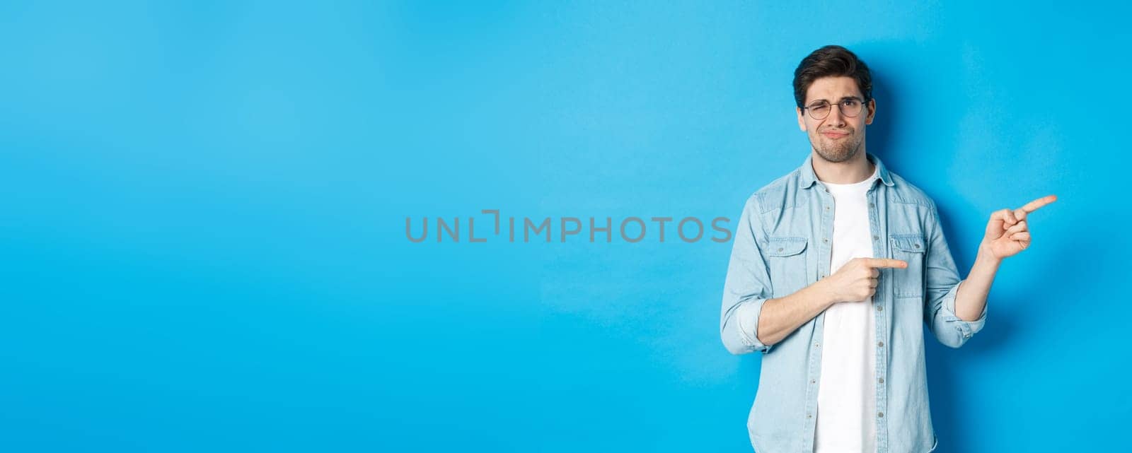 Disappointed young man in glasses pointing fingers right at copy space, showing promo banner and smirking dissatisfied, judging bad product, standing over blue background by Benzoix