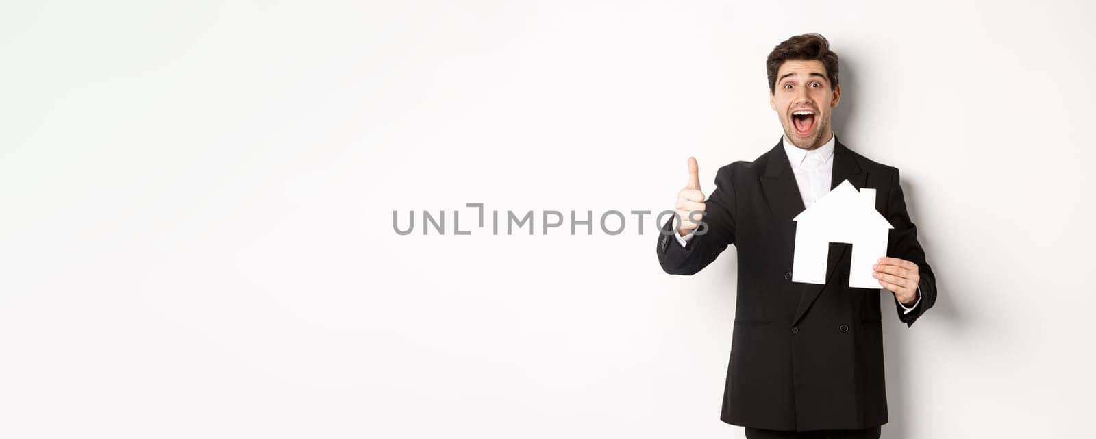 Portrait of excited handsome man looking for a home, showing house and thumb-up, recommending real estate agency, standing against white background.