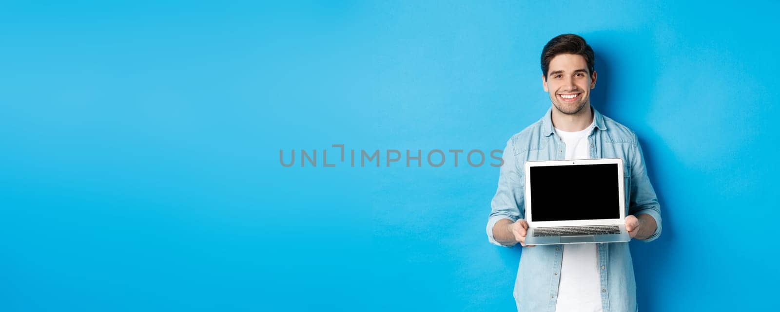 Handsome young man introduce product on laptop screen, showing computer and smiling, standing over blue background by Benzoix