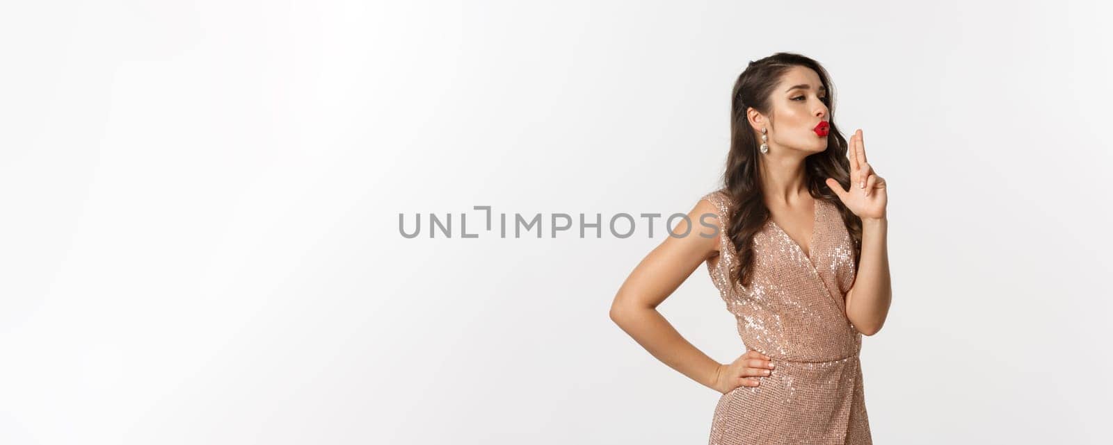 Concept of casino, celebration and party. Beautiful female model with red lips, wearing glamour dress, blowing at finger gun after making shot, standing over white background by Benzoix