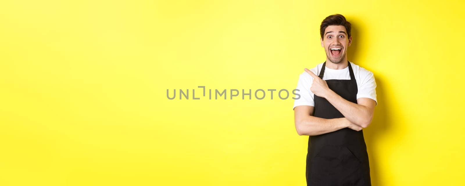 Surprised salesman in black apron pointing finger upper left corner, showing shop promo offer, standing against yellow background.
