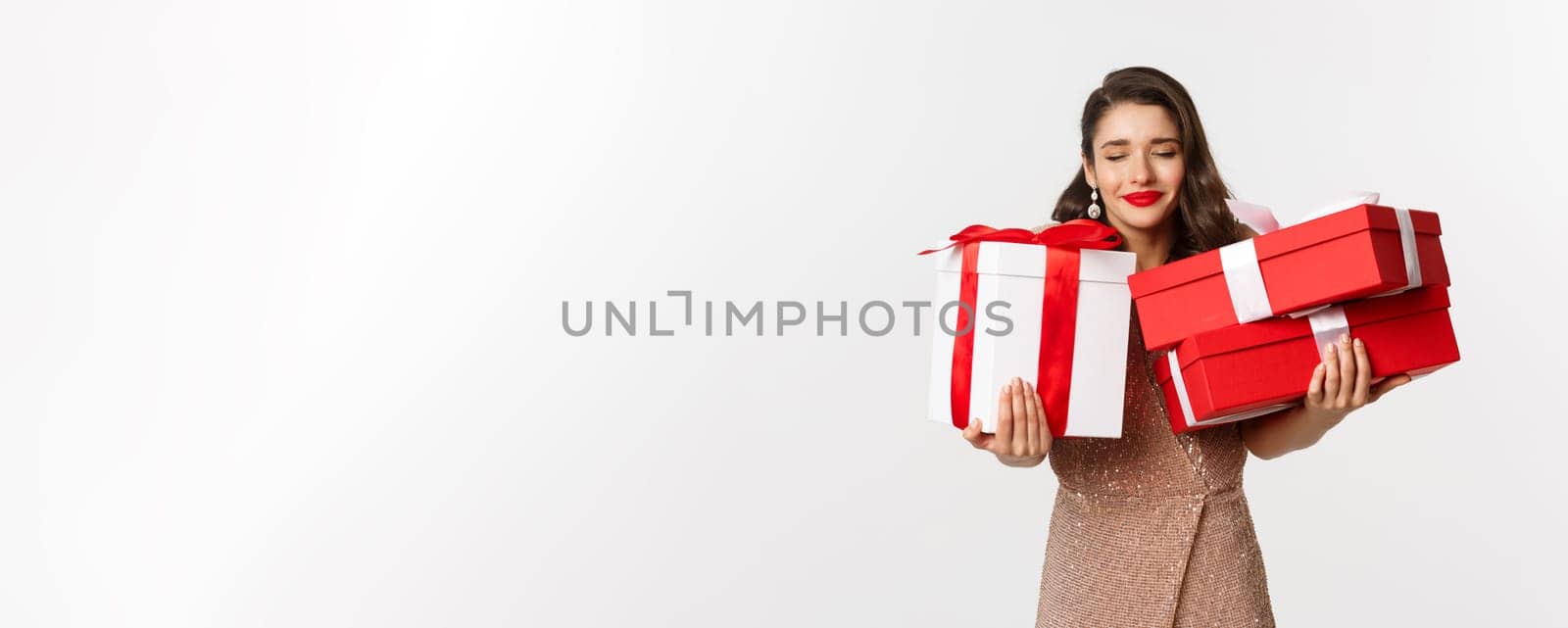 Holidays, celebration concept. Beautiful caucasian woman in elegant dress holding Christmas presents and smiling happy, standing over white background by Benzoix