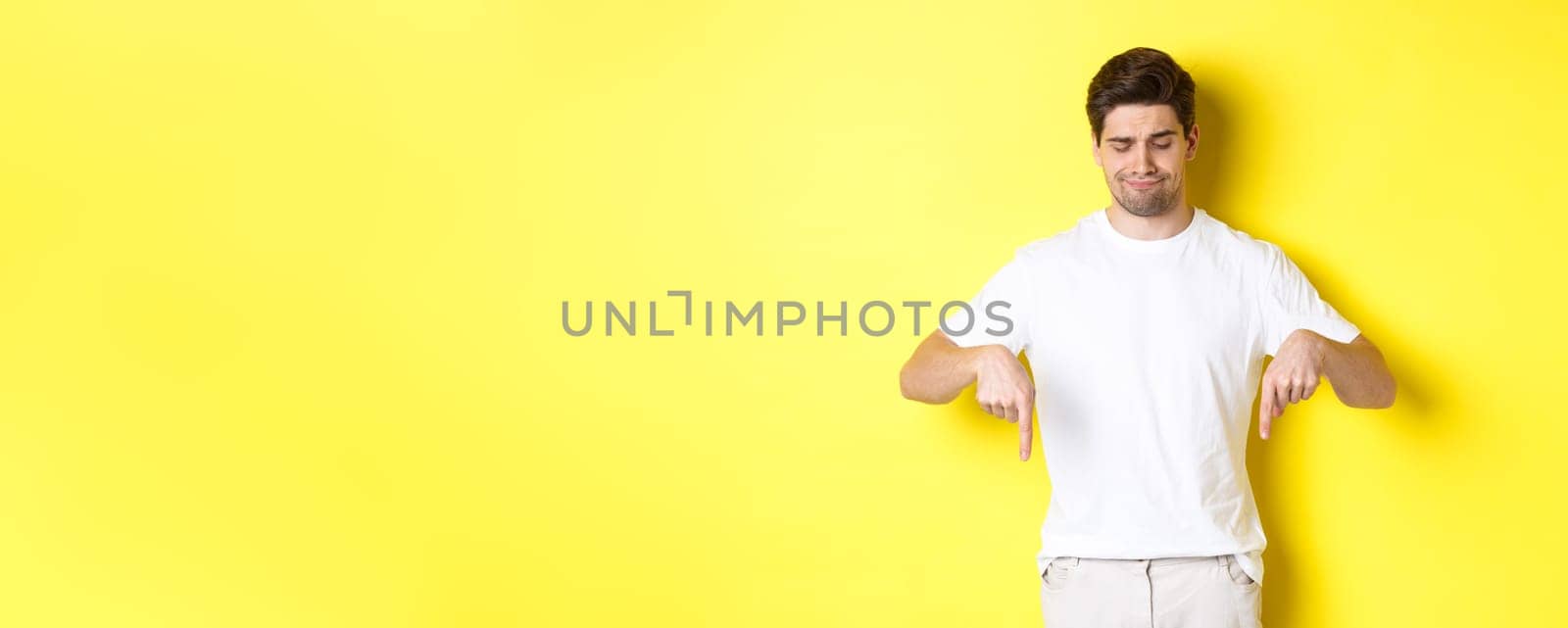 Skeptical young man in white t-shirt, pointing and looking down upset, disapprove and dislike product, standing over yellow background.