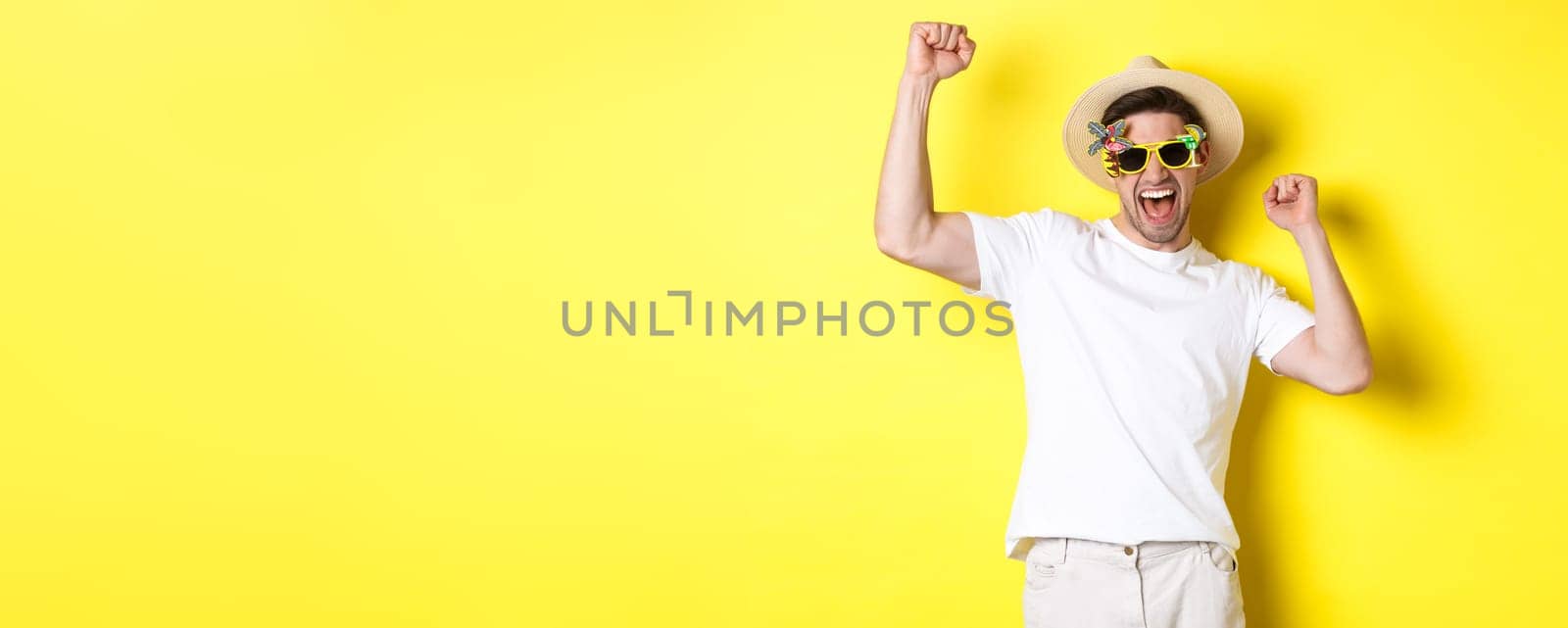 Concept of tourism and lifestyle. Happy lucky guy winning trip, rejoicing and wearing holiday outfit, summer hat and sunglasses, yellow background.