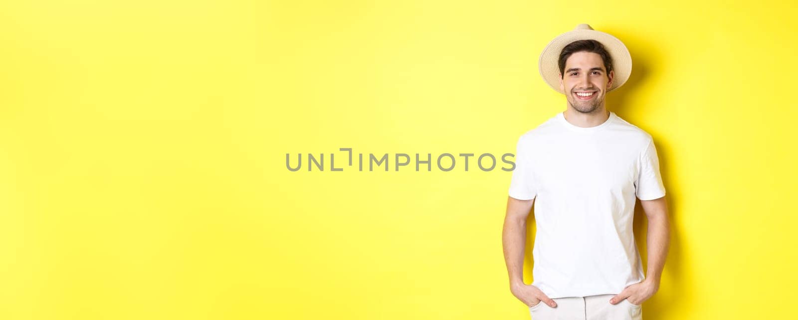 Young handsome guy tourist looking happy, wearing straw hat for travelling, standing against yellow background. Copy space