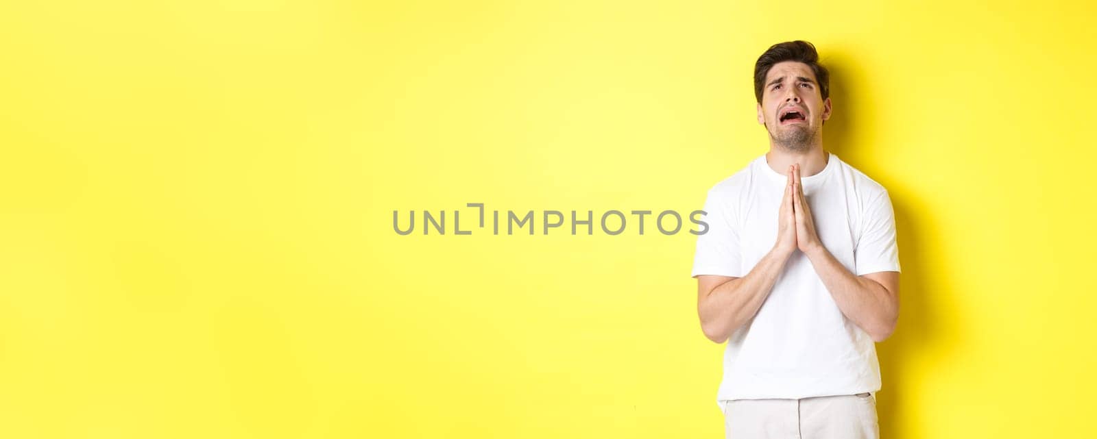 Desperate man pleading God, holding hands in pray and looking up distressed, standing over yellow background. Copy space