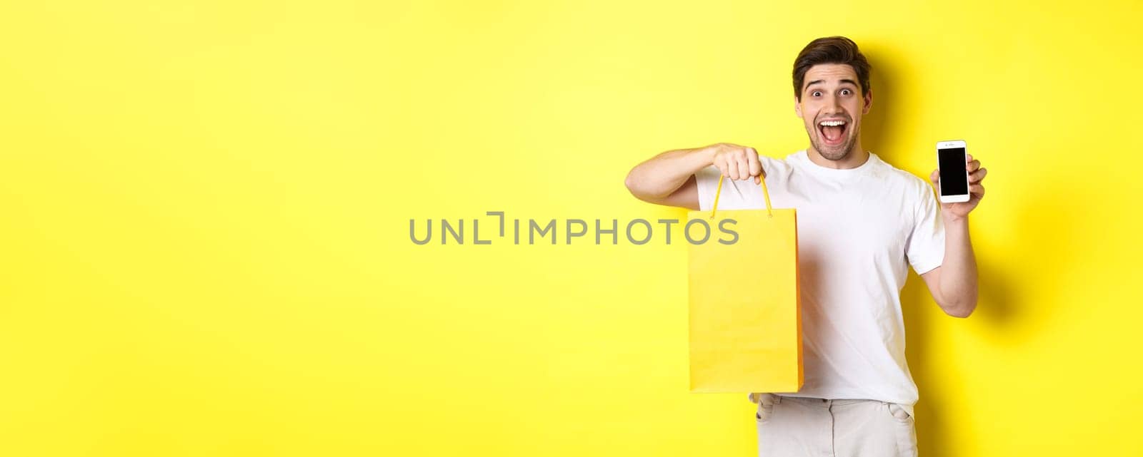 Concept of mobile banking and cashback. Young happy guy holding shopping bag and showing smartphone screen, yellow background by Benzoix
