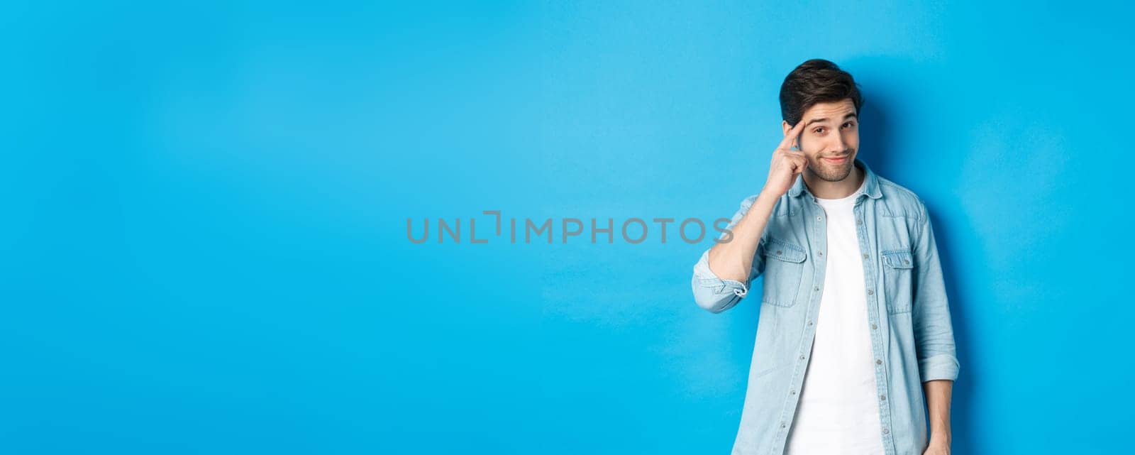 Young thoughtful man pointing at head, asking to think about it, giving a hint, standing in casual clothes over blue background.