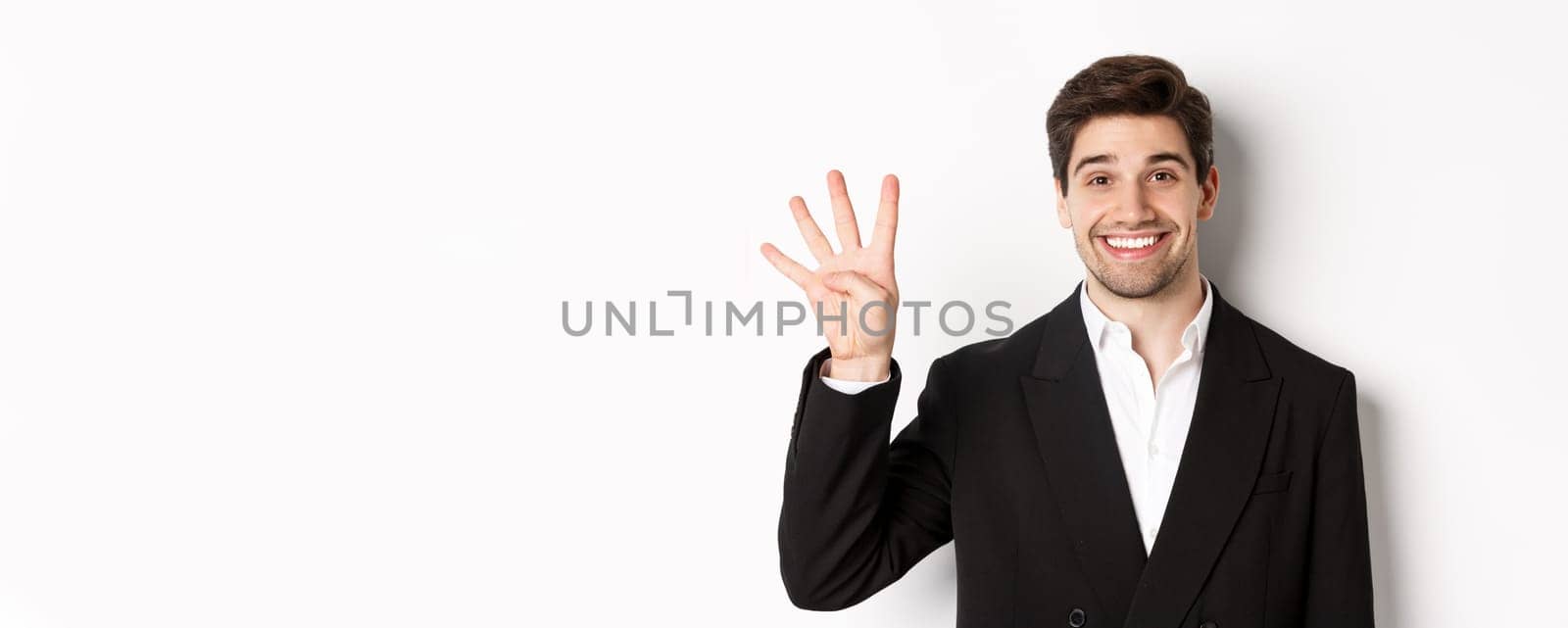 Close-up of handsome businessman in black suit, smiling amazed, showing number four, standing over white background.