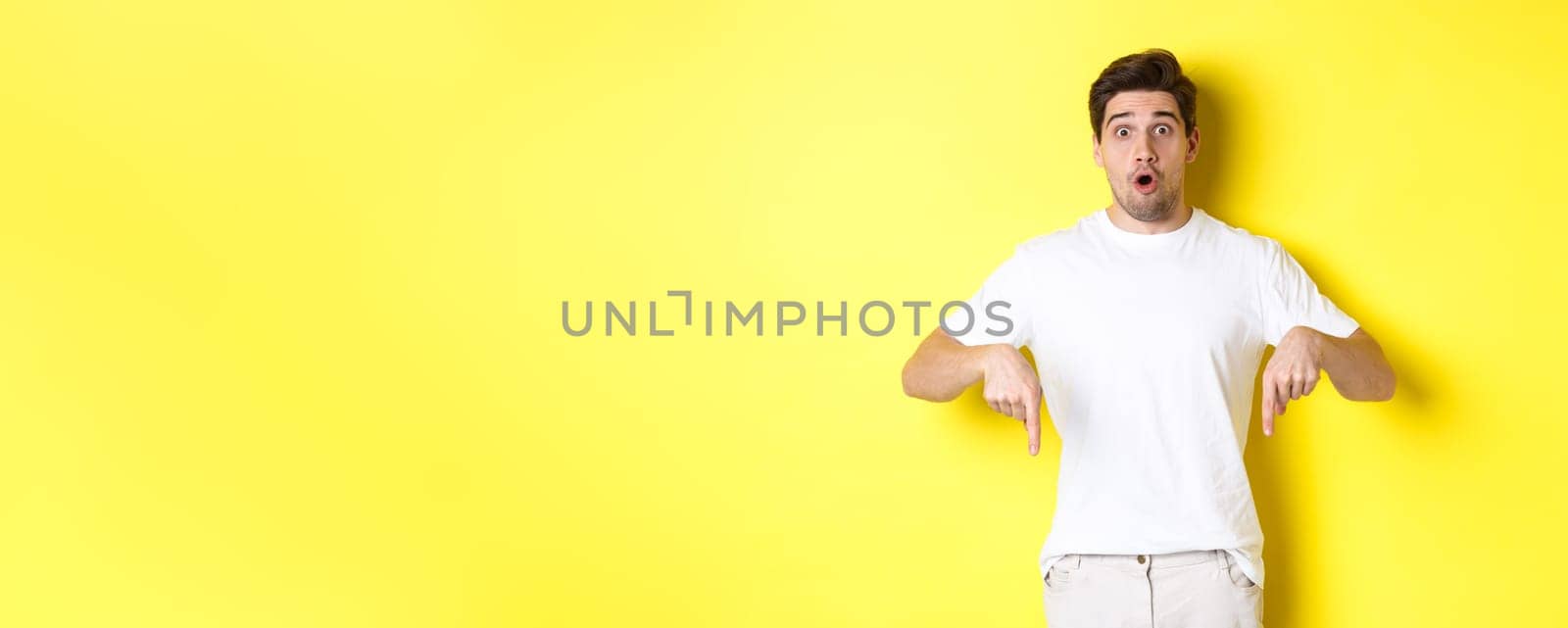Image of impressed guy pointing fingers down, gasping amazed and looking at camera, standing over yellow background.