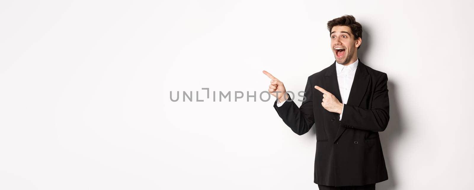 Portrait of amazed and happy handsome man in black suit, pointing and looking left impressed, showing awesome christmas promo, standing over white background.