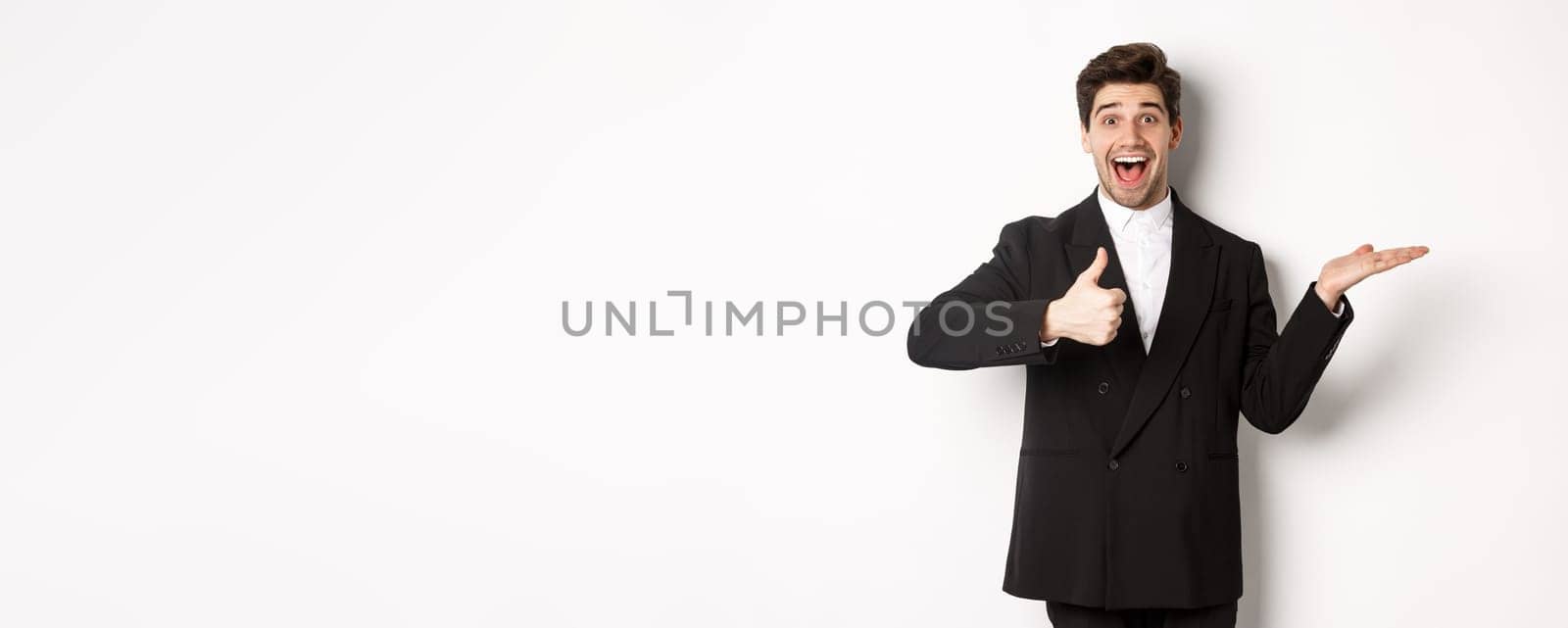 Portrait of hadnsome bearded man in formal suit, showing thumb-up and holding product in hand over white copy space, recommending product, standing over white background.