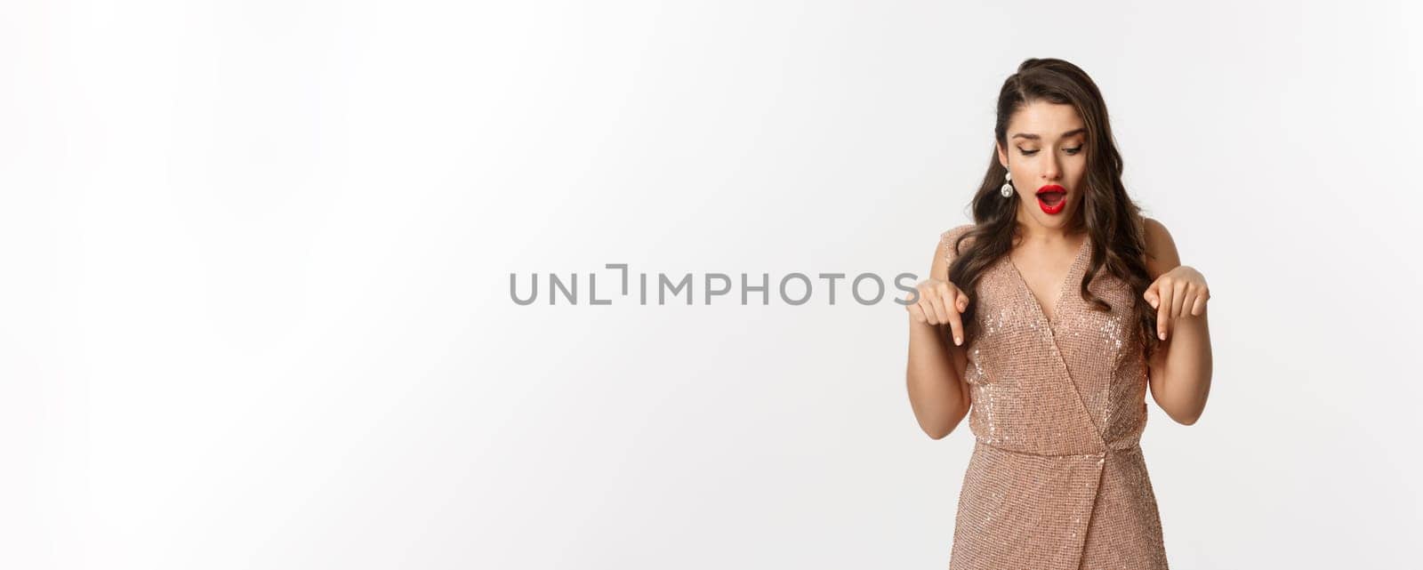 Amazed glamour girl in party dress, looking and pointing down amazed, showing christmas advertisement, standing over white background.