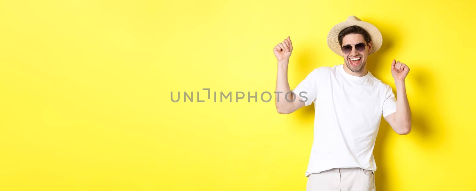 Tourism, travelling and holidays concept. Happy caucasian guy dancing and having fun on vacation, wearing sunglasses with straw hat, standing against yellow background.