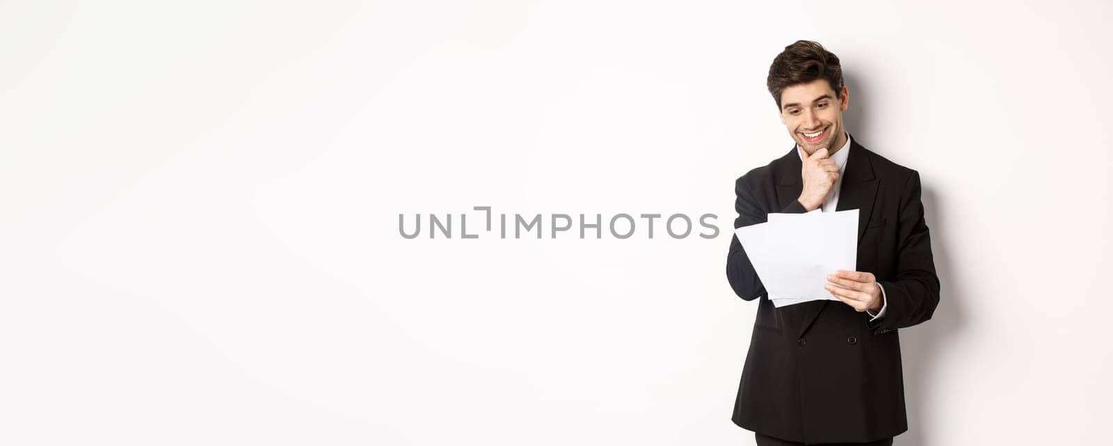 Image of handsome businessman in black suit, looking pleased at documents, reading report and smiling, standing against white background.