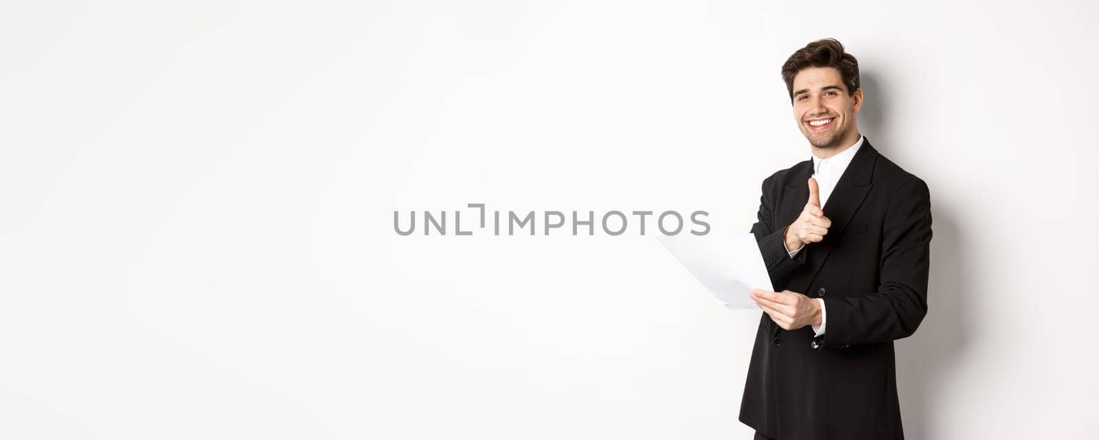 Image of handsome businessman in black suit, holding document and pointing finger at camera, praising good job, standing against white background.