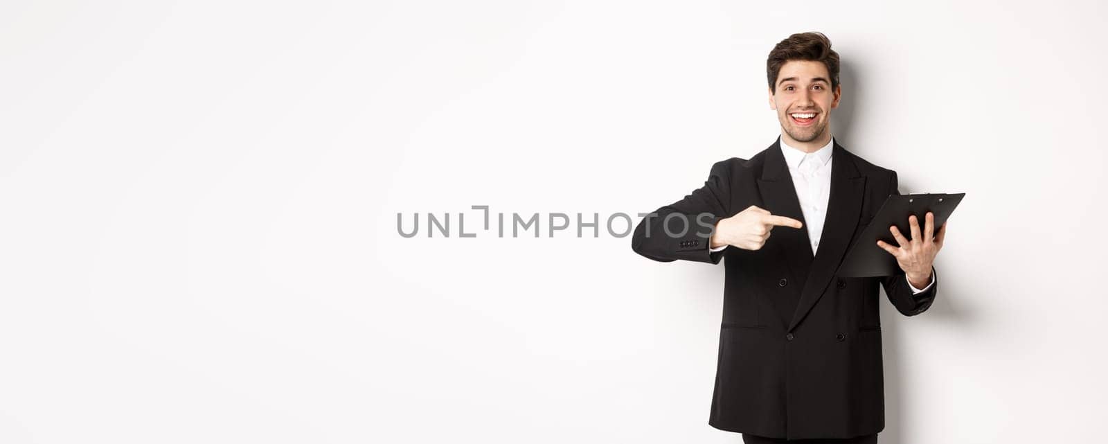 Image of handsome smiling businessman in black suit, pointing finger at clipboard with documents, standing against white background by Benzoix
