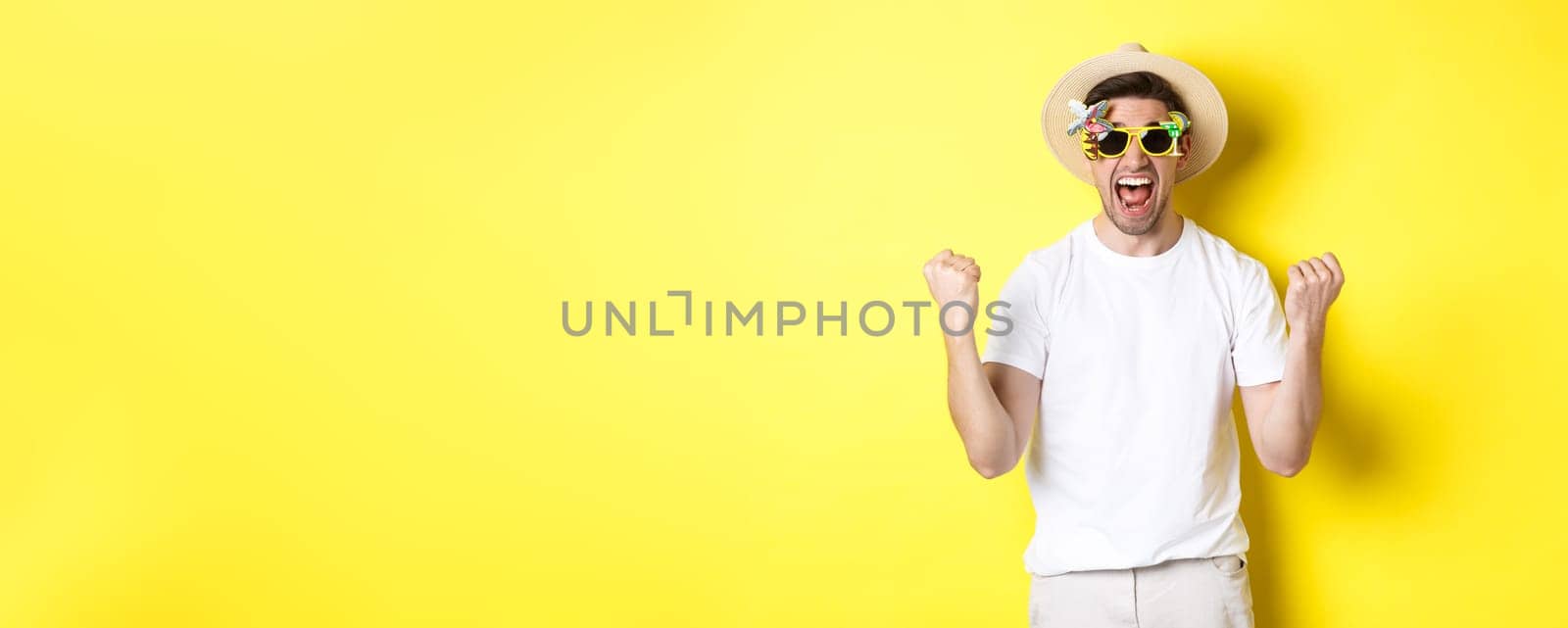 Happy guy going on vacation, winning or celebrating, wearing summer hat and sunglasses. Tourist looking excited, standing against yellow background by Benzoix