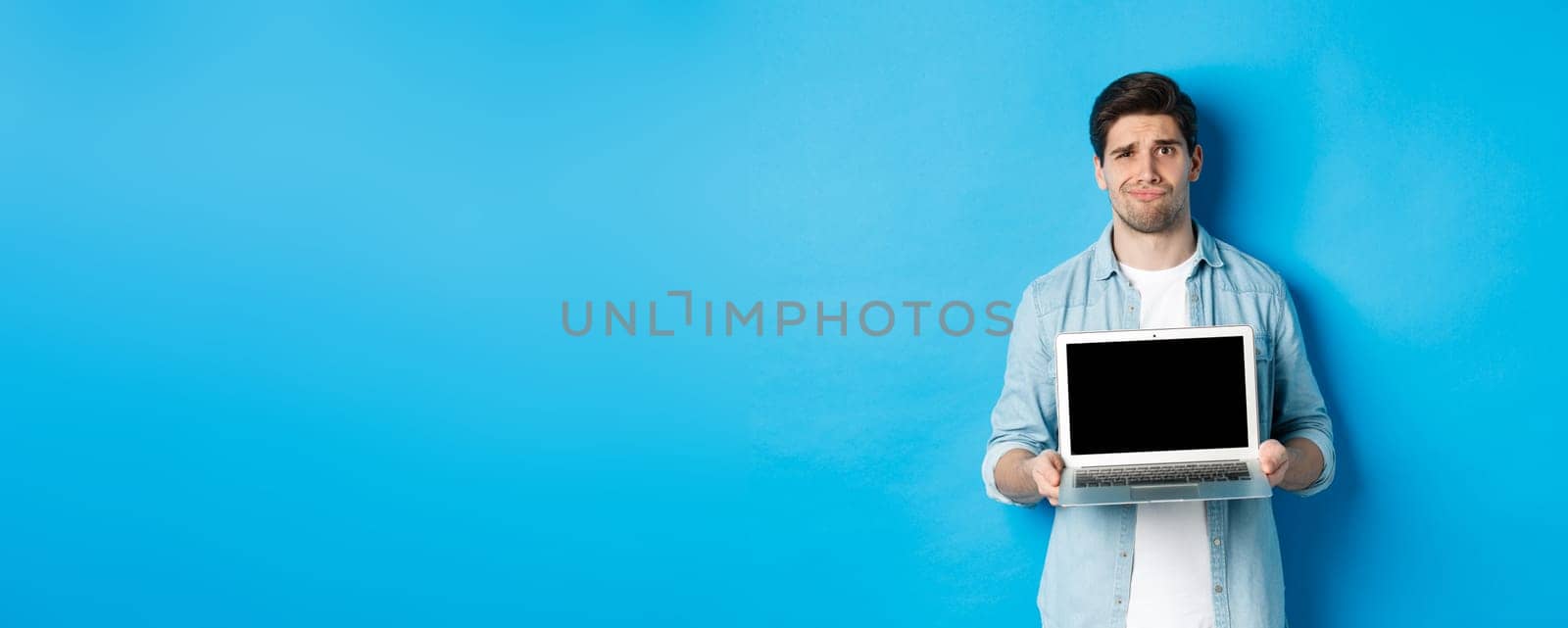 Skeptical and displeased bearded guy showing laptop screen and grimacing, having doubts, standing over blue background in casual clothes by Benzoix