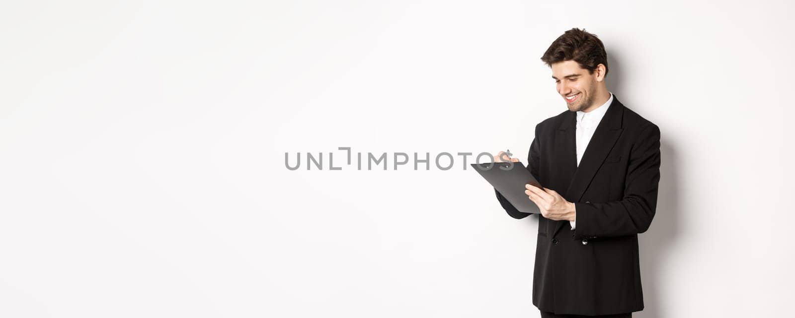 Image of handsome businessman in suit signing documents, looking at clipboard and smiling, standing against white background by Benzoix