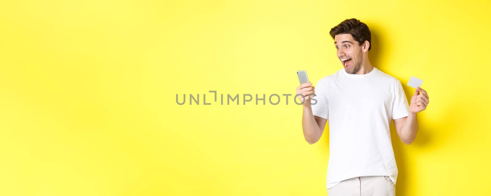 Surprised guy holding smartphone and credit card, online shopping on black friday, standing over yellow background by Benzoix