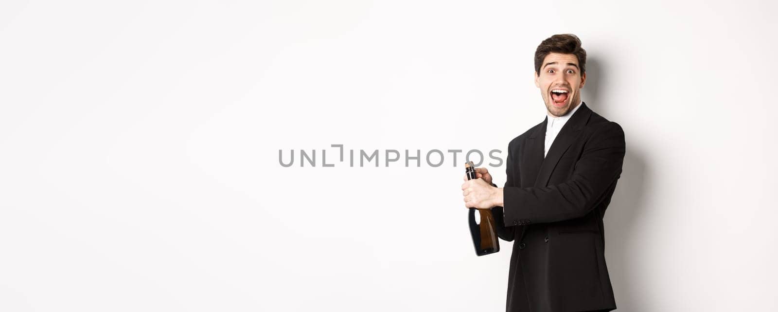 Portrait of attractive man in black suit, winking at camera and opening bottle of champagne, celebrating new year, standing against white background.
