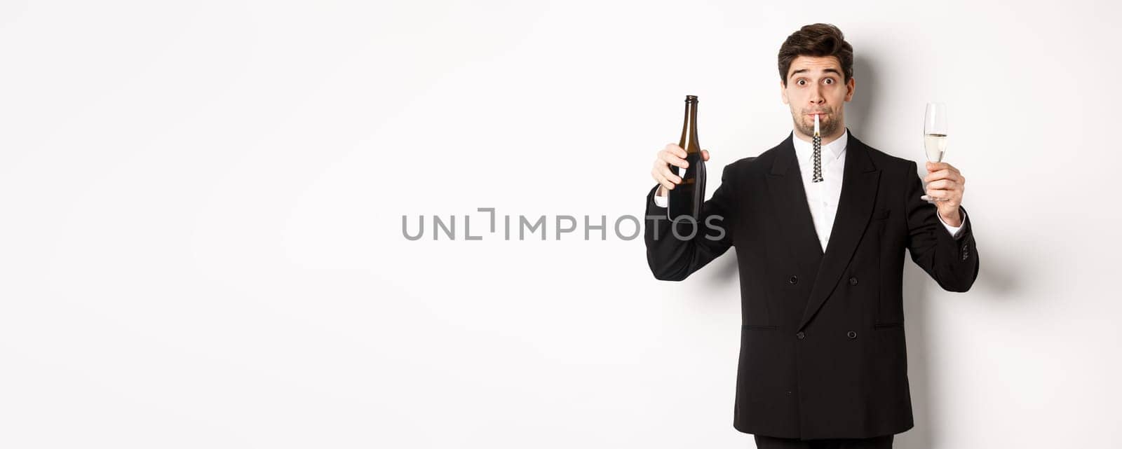 Concept of holidays, party and celebration. Portrait of handsome guy in black suit, raising bottle of champagne and glass, blowing a party whistle, having a birthday, standing over white background by Benzoix