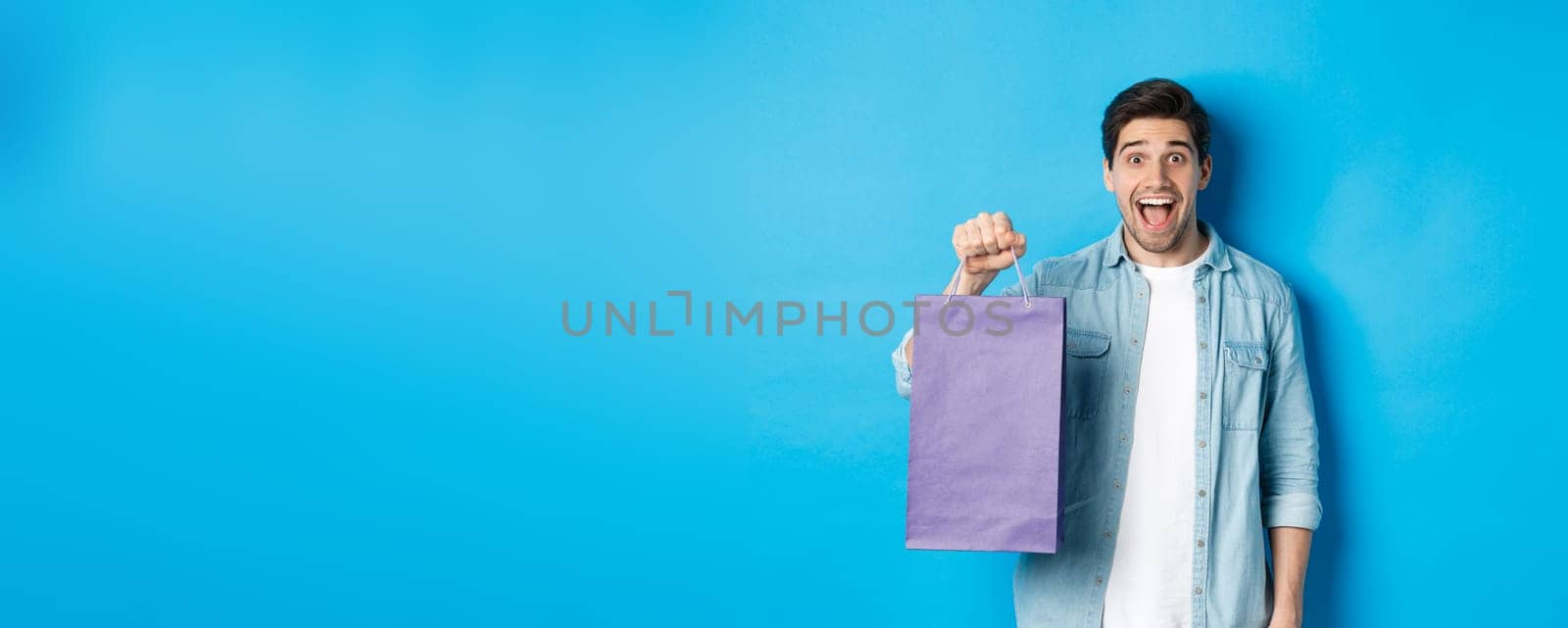 Concept of shopping, holidays and lifestyle. Excited handsome guy holding paper bag with gift and looking happy, standing over blue background by Benzoix