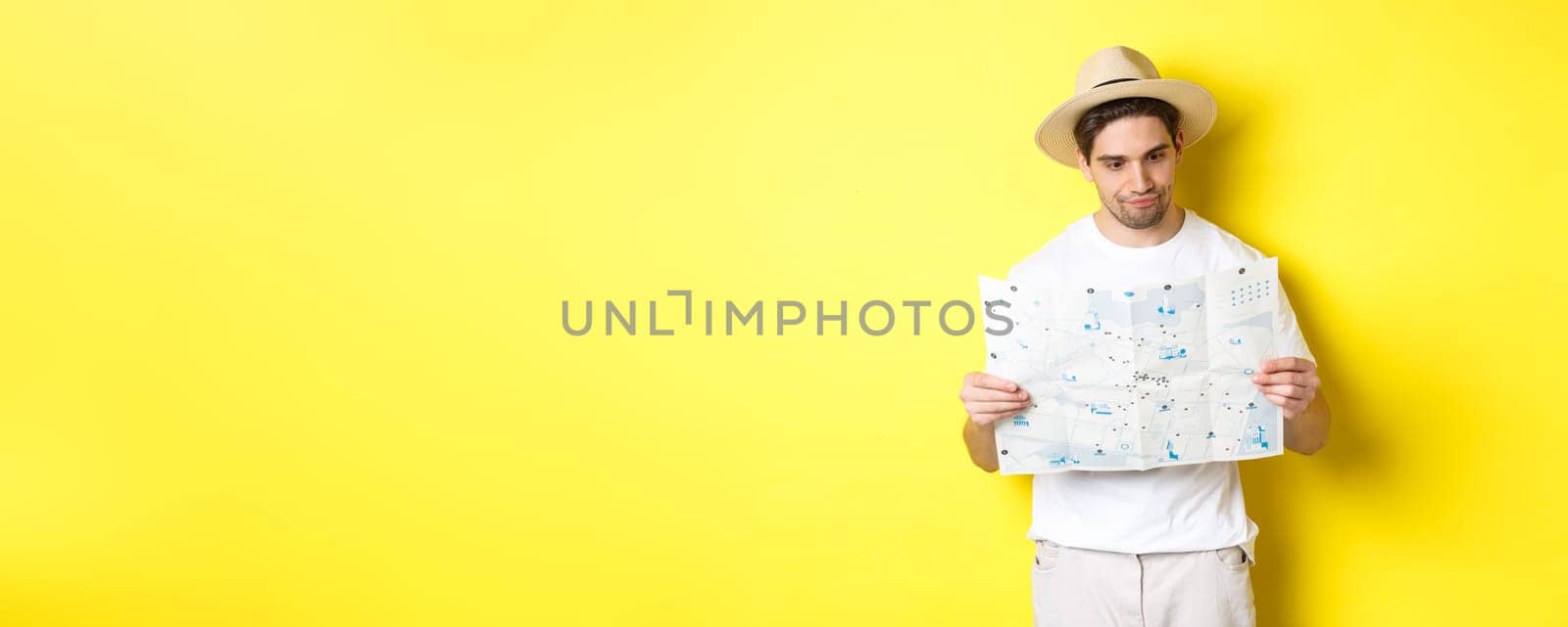 Travelling, vacation and tourism concept. Male tourist looking confused at map, looking for right direction, standing over yellow background.