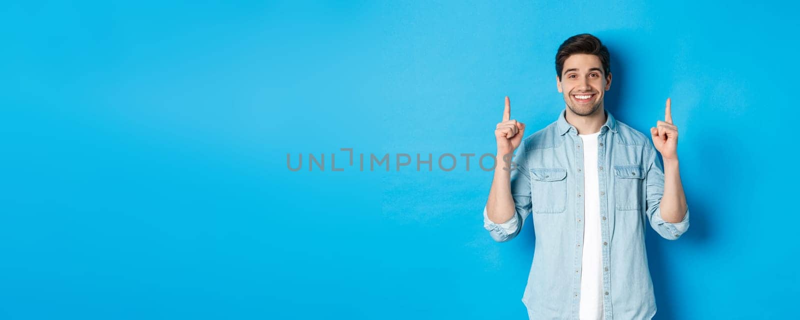 Portrait of handsome bearded man in casual outfit, smiling happy and pointing fingers up at copy space, standing over blue background by Benzoix